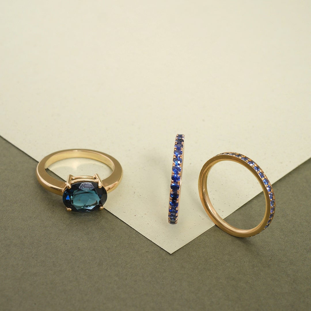 Anillos en oro con zafiros azules y topacio London, diseño artesanal de Mariana Shuk sobre fondo en tonos gris y crema.