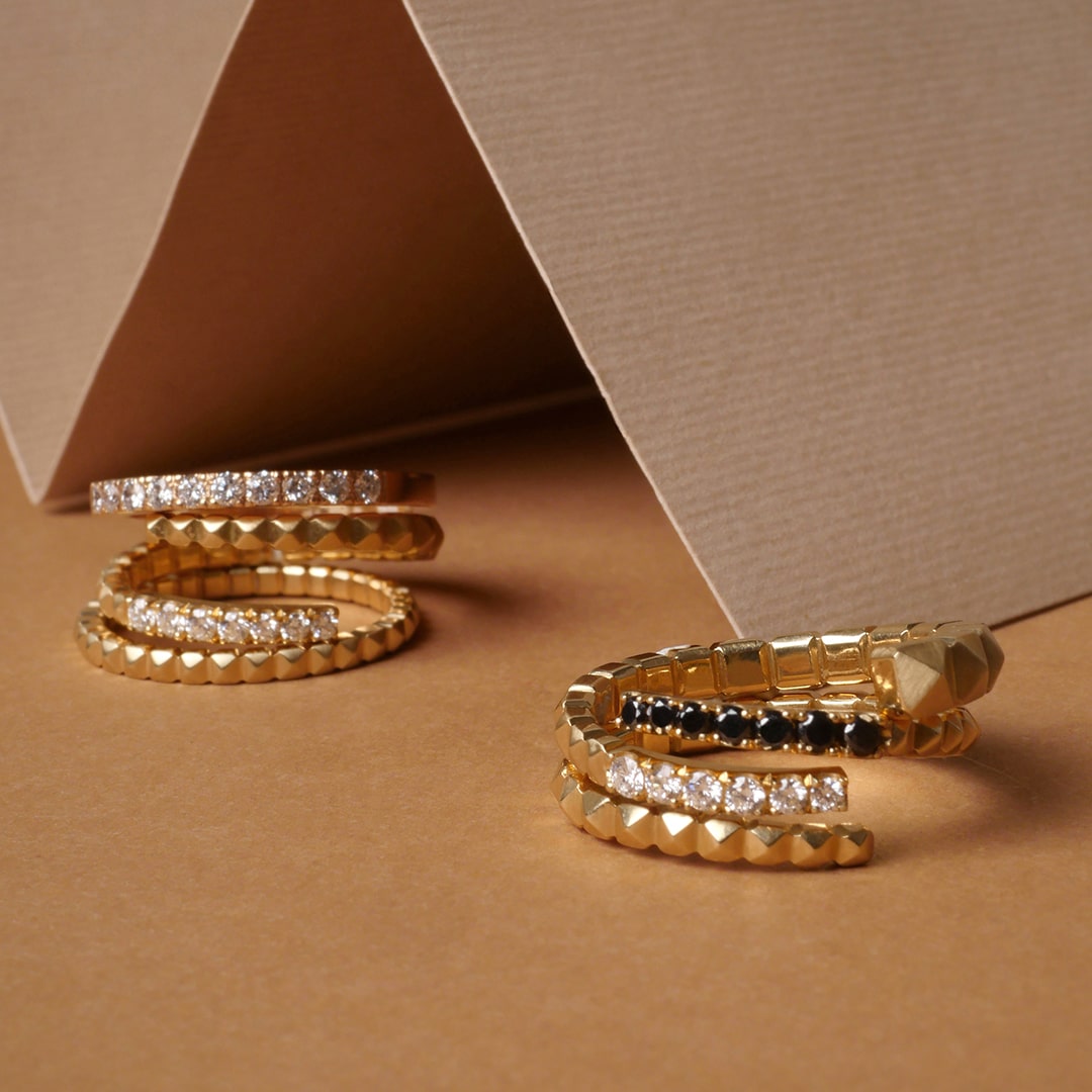 Anillos en oro con diamantes y ónix negro, diseños tipo serpiente y caracol de Mariana Shuk sobre fondo beige con textura artesanal.