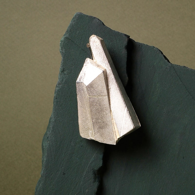Broche en plata 925 con diseño geométrico facetado, pieza escultórica artesanal de Mariana Shuk sobre fondo de piedra verde oscura.