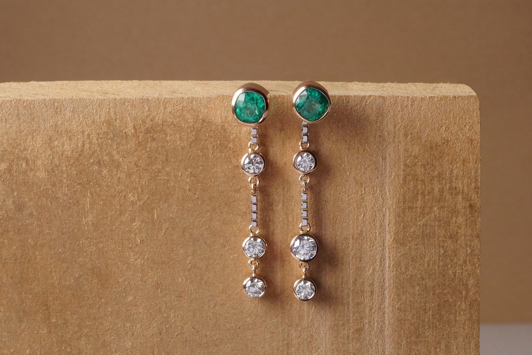 Aretes largos en oro con esmeraldas cushion colombianas y seis diamantes talla brillante, diseño artesanal de Mariana Shuk sobre fondo de madera minimalista.