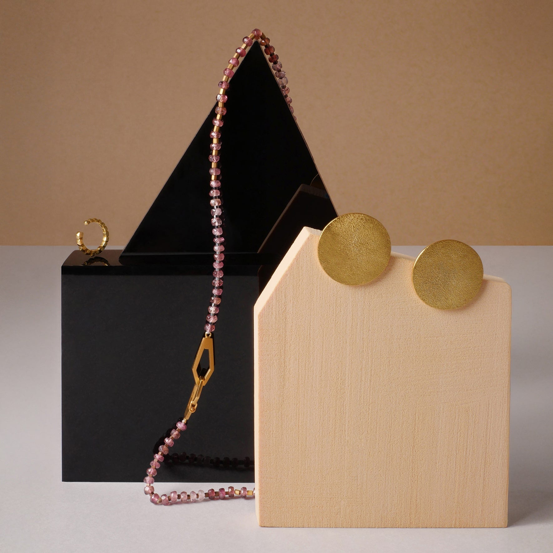 Aretes circulares dorados, collar de turmalinas con dije geométrico y orejera de taches en oro, diseño artesanal de Mariana Shuk sobre bloques de madera y acrílico negro.