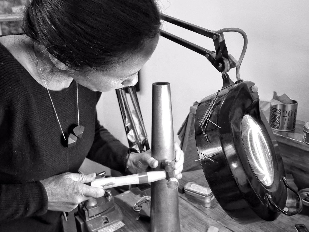 La joyera colombiana Mariana Shuk trabajando con precisión bajo una lámpara de aumento en su taller de joyería en Bogotá. Usa el collar de dos poliedros.