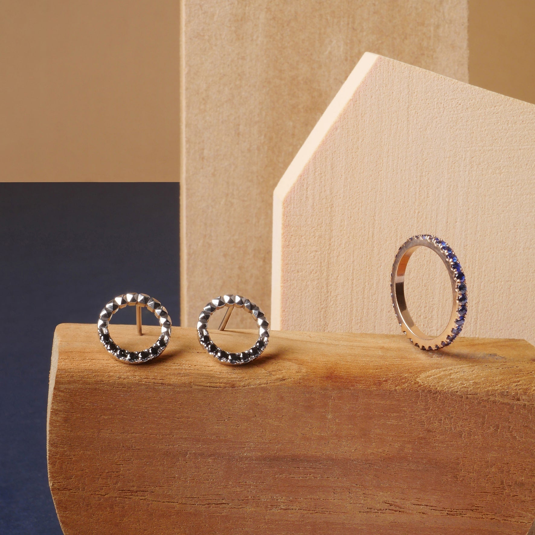 Aretes tipo taches en oro con diamantes negros y argolla sin fin con zafiros azules, joyas de diseño artesanal Mariana Shuk sobre fondo de madera clara.