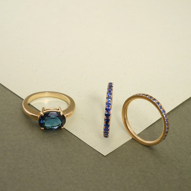 Anillos en oro con zafiros azules y topacio London, diseño artesanal de Mariana Shuk sobre fondo en tonos gris y crema.