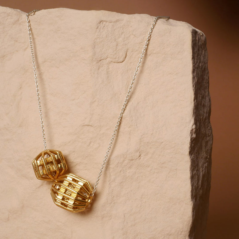 Collar con cadena de plata y dijes geométricos dorados tipo globo, diseño artesanal de Mariana Shuk sobre piedra color arena.