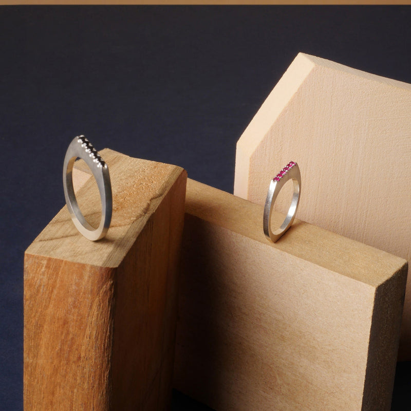Anillos planos en plata 925 con incrustaciones de rubíes y diamantes negros, diseño contemporáneo de Mariana Shuk sobre bloques de madera.