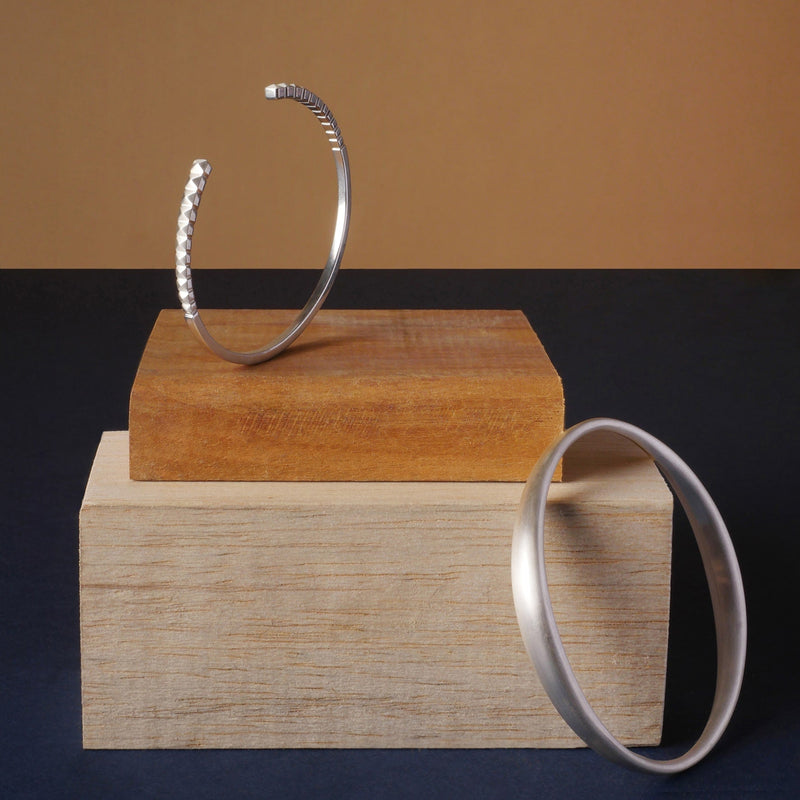 Brazaletes en plata 925, uno con diseño facetado tipo taches y otro liso, joyería artesanal de Mariana Shuk sobre bloques de madera.