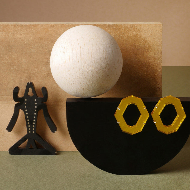 Aretes hexagonales en acrílico amarillo con herrajes en plata 925, diseño artístico de Mariana Shuk inspirados en geometría contemporánea, sobre fondo de madera y figuras escultóricas.