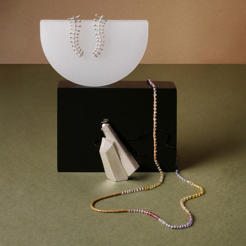 Collar de zafiros multicolor con dije geométrico, broche de cuarzo y aretes en plata con diseño de líneas y puntos, piezas artesanales de Mariana Shuk sobre fondo verde y beige.