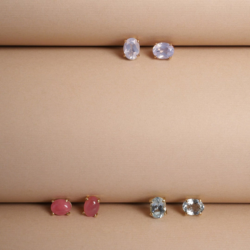 Aretes en oro con piedras semipreciosas —piedra luna, aguamarina y rodocrosita— diseño artesanal de Mariana Shuk sobre fondo beige texturizado.