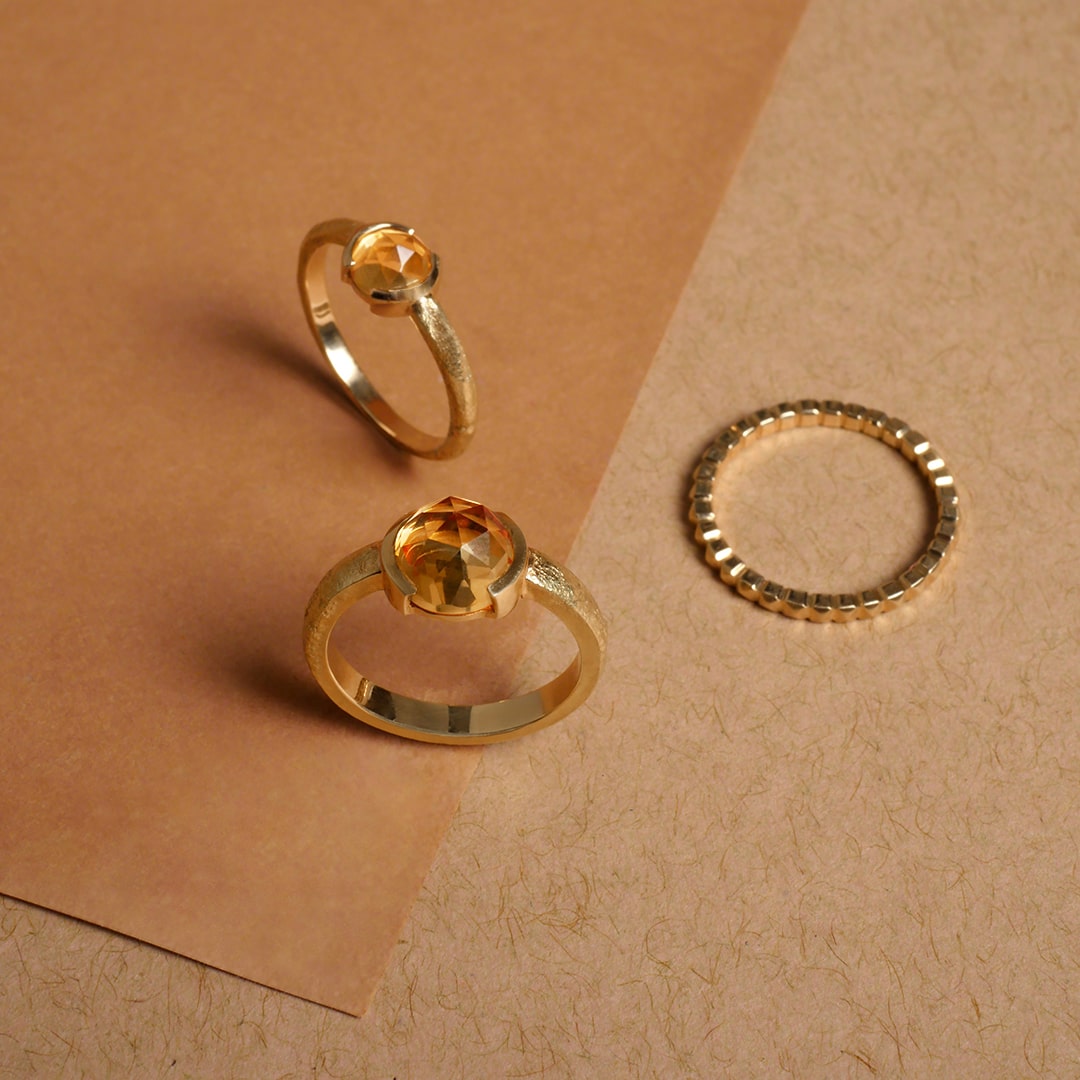 Anillos en oro con piedra citrino facetada y argolla texturizada, diseño artesanal de Mariana Shuk sobre fondo en tonos tierra.