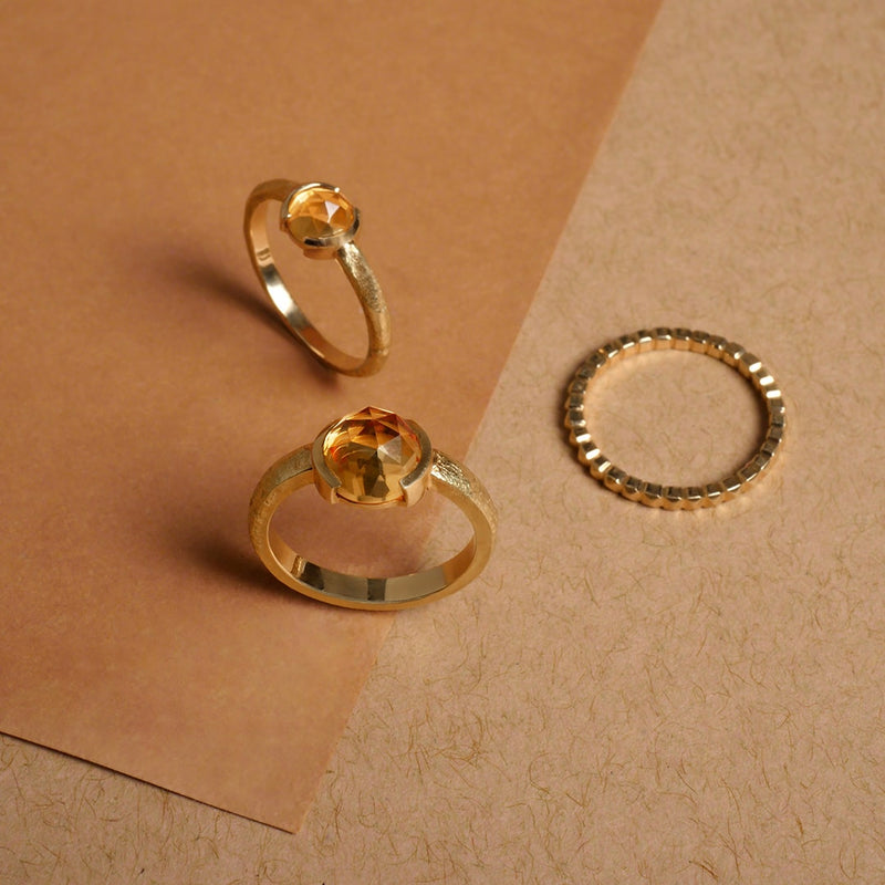 Anillos en oro con piedra citrino facetada y argolla texturizada, diseño artesanal de Mariana Shuk sobre fondo en tonos tierra.