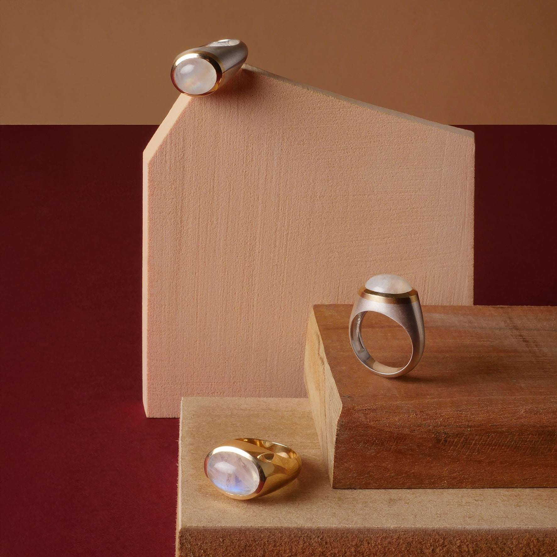 Anillos en plata y oro con piedra luna, diseño artesanal de Mariana Shuk sobre bloques de madera y fondo rojo terracota. Mariana Shuk joyería de autor.