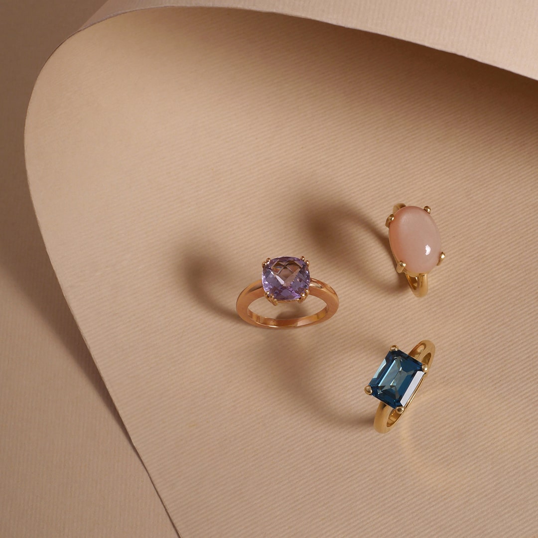 Anillos en oro amarillo con amatista, cuarzo rosa y topacio azul, joyería artesanal de Mariana Shuk sobre fondo beige texturizado.