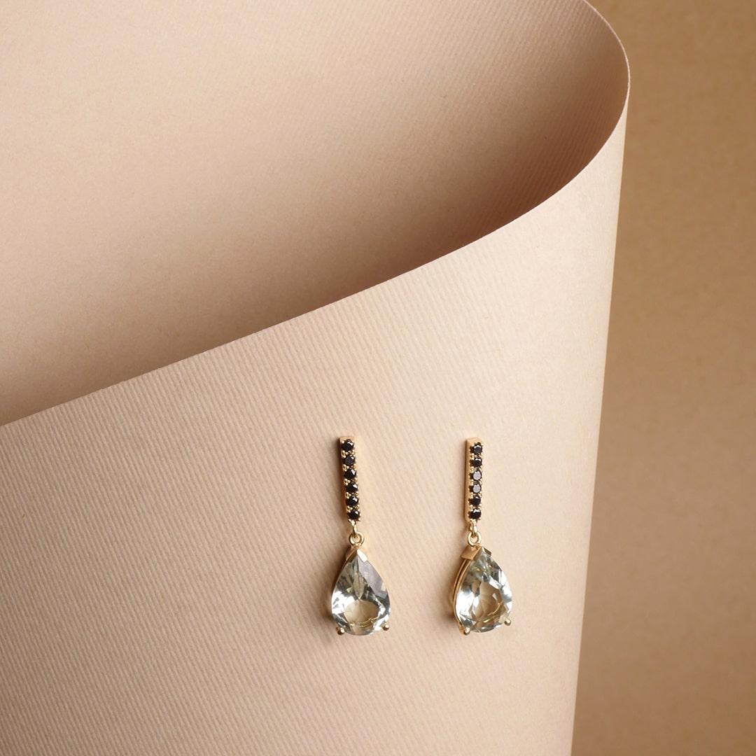 Aretes en oro con diamantes negros y cuarzo verde en forma de gota, diseño artesanal de Mariana Shuk sobre fondo beige texturizado.