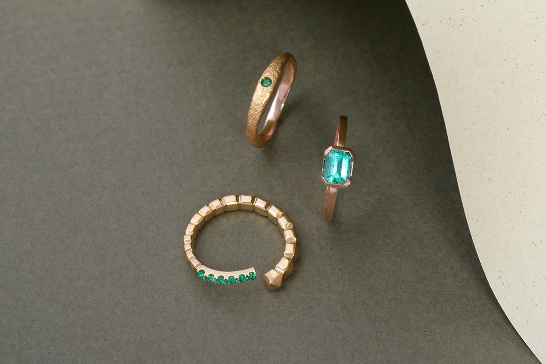 Tres anillos en oro con esmeraldas colombianas, diseño contemporáneo de Mariana Shuk sobre fondo gris con textura y papel curvado. Formato banner. Alta joyería.