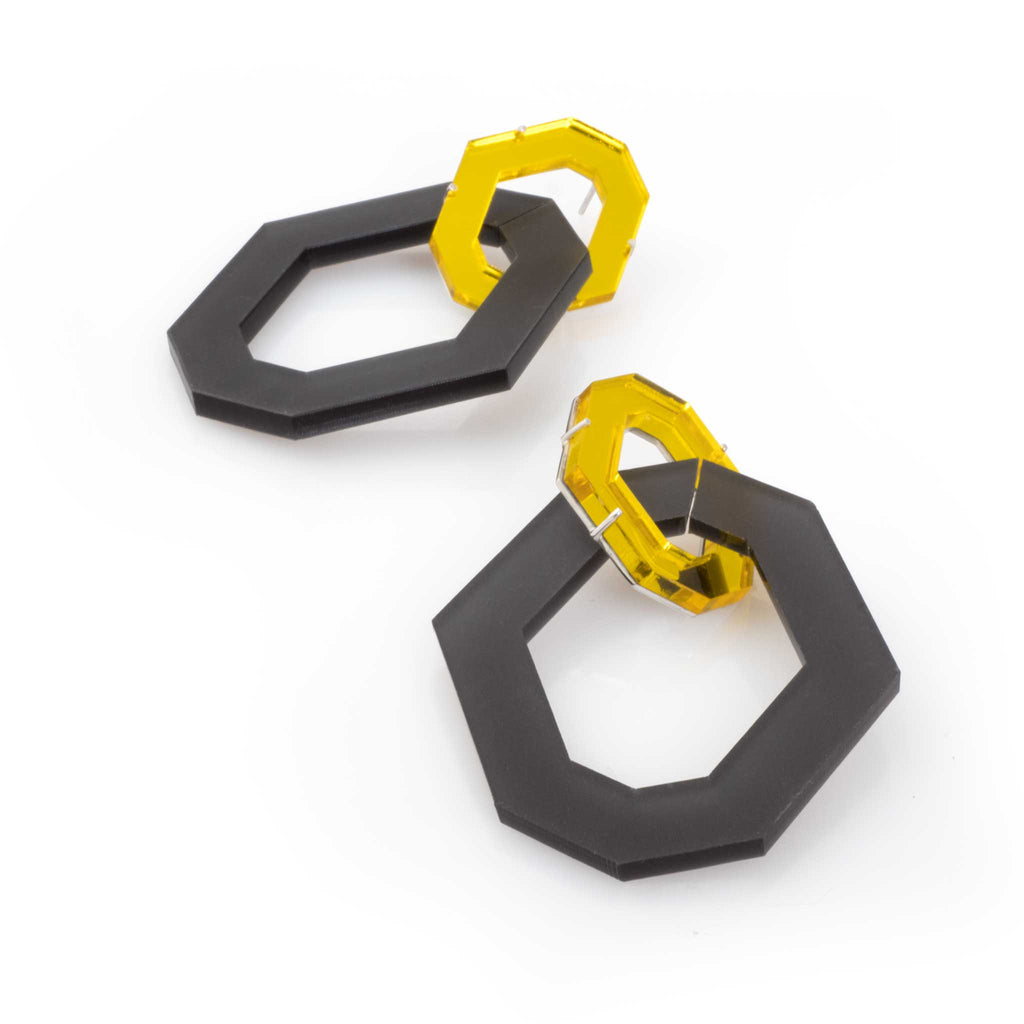 Aretes dos hexágonos en acrílico esmerilado negro y acrílico espejado amarillo, foto con vista frontal. Joyería artesanal inspirada en contraste y simetría. Diseños exclusivos, hecho en Colombia por Mariana Shuk.